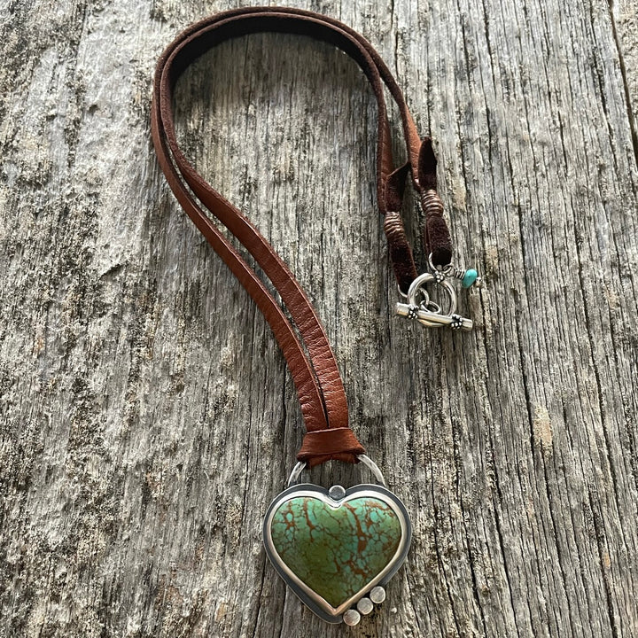 Green and brown matrix Kingman turquoise heart set in sterling silver with three silver accent discs, on a brown leather cord, displayed on weathered wood.