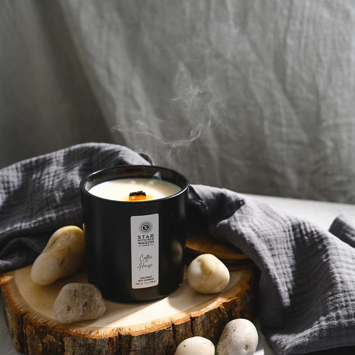 Coffee House Candle with wooden wick just blown out in a matte black jar with label. Linen towel, rocks and wood log surround candle.