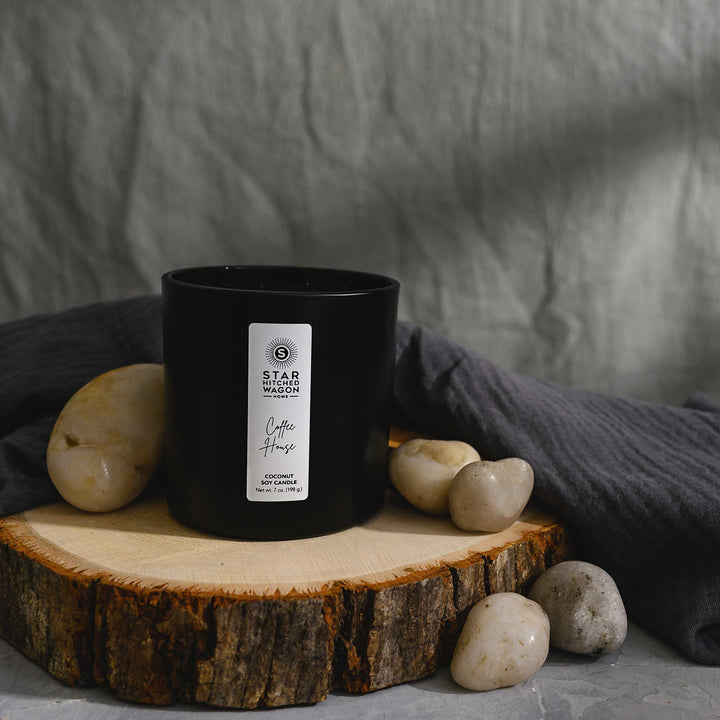 Coffee House Candle in a matte black jar with label. Linen towel, rocks and wood log surround candle.