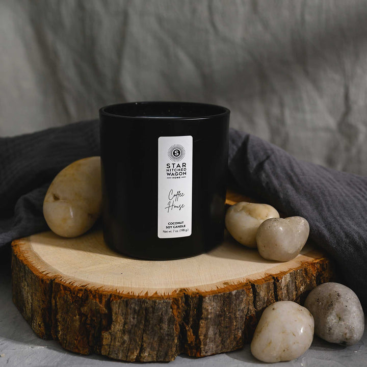 Coffee House Candle in a matte black jar with label. Linen towel, rocks and wood log surround candle.