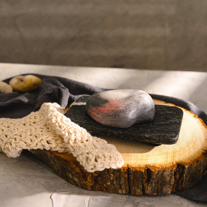 Margo river stone salt soap bar, placed on a wooden slice next to a luffa, with a dark cloth in the background.