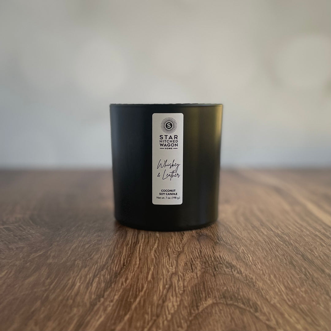whiskey and leather candle in matte black container with label, off white and wood texture background