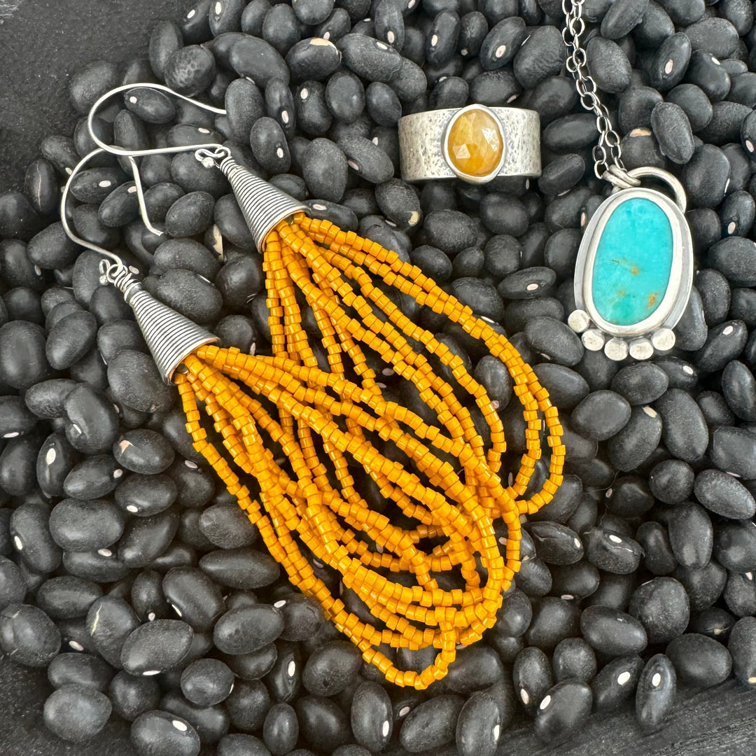 Mustard yellow seed bead earrings, a hammered silver and yellow sapphire ring, and a turquoise pendant necklace displayed on black beans. 