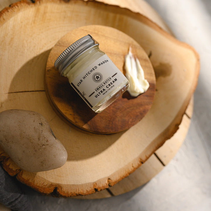 A small glass jar of Star Hitched Wagon Nora Ultra Cream lays on a round wooden slice, with a dollop of ultra cream next to jar.