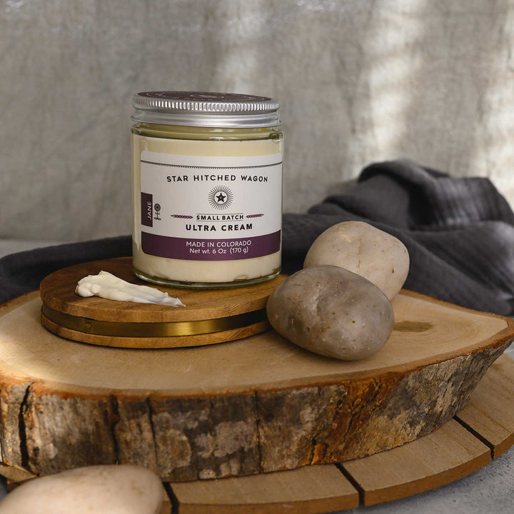 Jane Ultra Cream in a glass jar placed on a wooden disc, alongside some stones and a grey towel. 