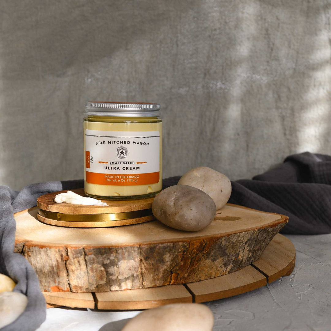 Jess Ultra Cream in a glass jar placed on a wooden platform, pebbles and grey towel in foreground and background.