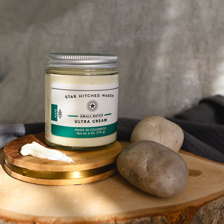 Kate Ultra Cream in a glass jar with an aluminum screw top lid, displayed on a wooden surface with a grey cloth and pebbles in the background.