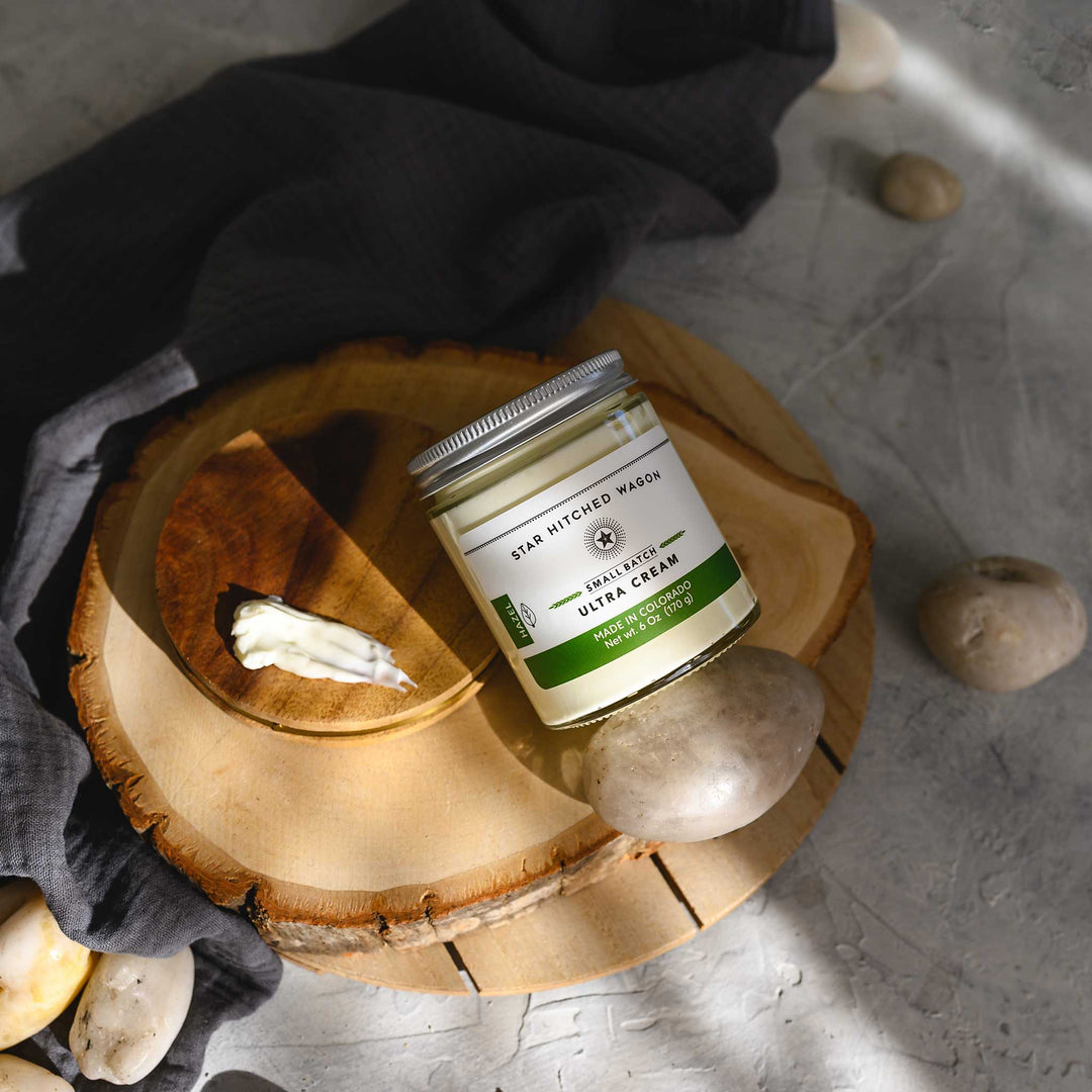 Hazel Ultra Cream in a glass jar with an aluminum screw top lid, placed on a wooden surface with pebbles and grey towel in the background.