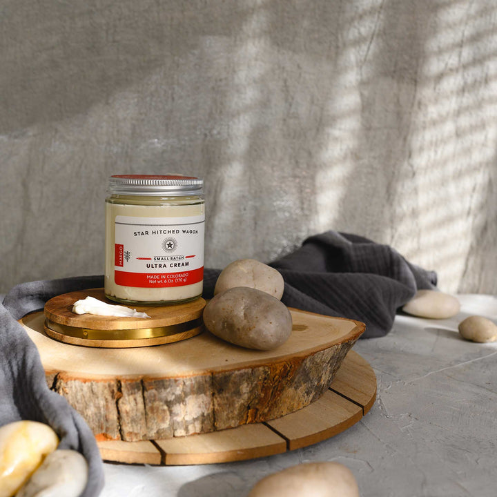 Margo Ultra Cream in a glass jar with an aluminum screw top lid, displayed on a wooden surface with a grey cloth and pebbles in the background.
