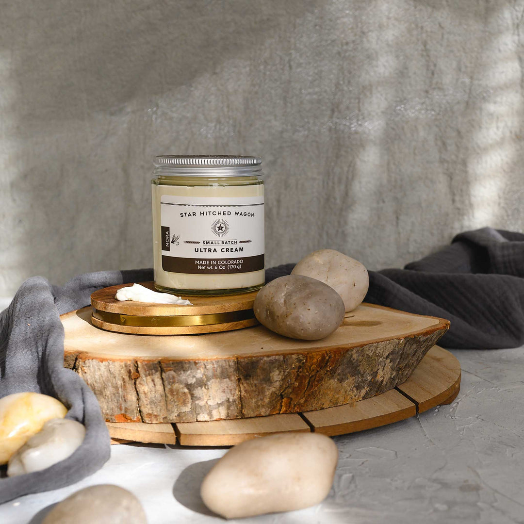 Nora Ultra Cream in a glass jar with an aluminum screw top lid, displayed on a wooden surface with a grey cloth and pebbles in the background.