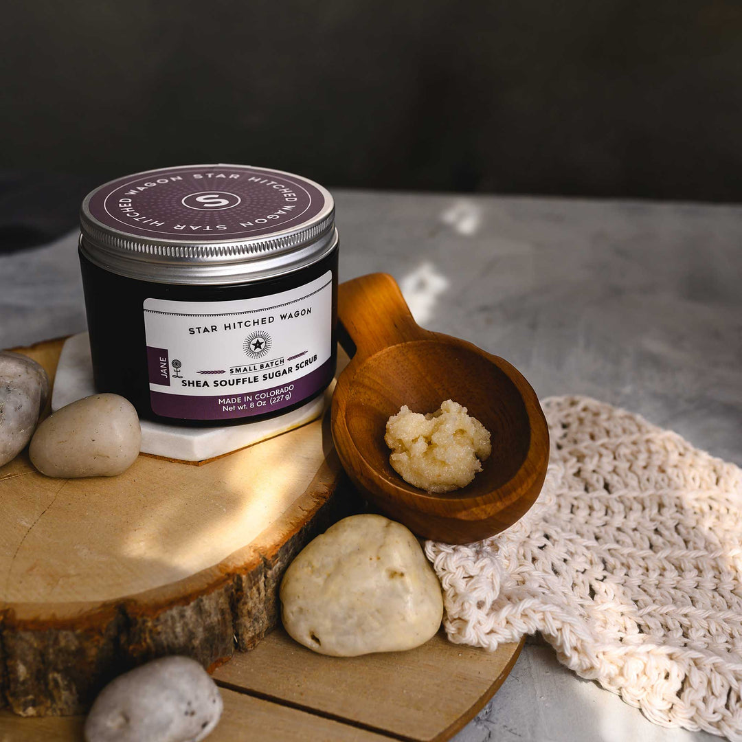 A jar of Jane 8 Ounce Shea Soufflé Sugar Scrub is displayed on a wooden slice, alongside a wooden scoop and a knit washcloth, with a few stones around. The setup is on a marble surface.