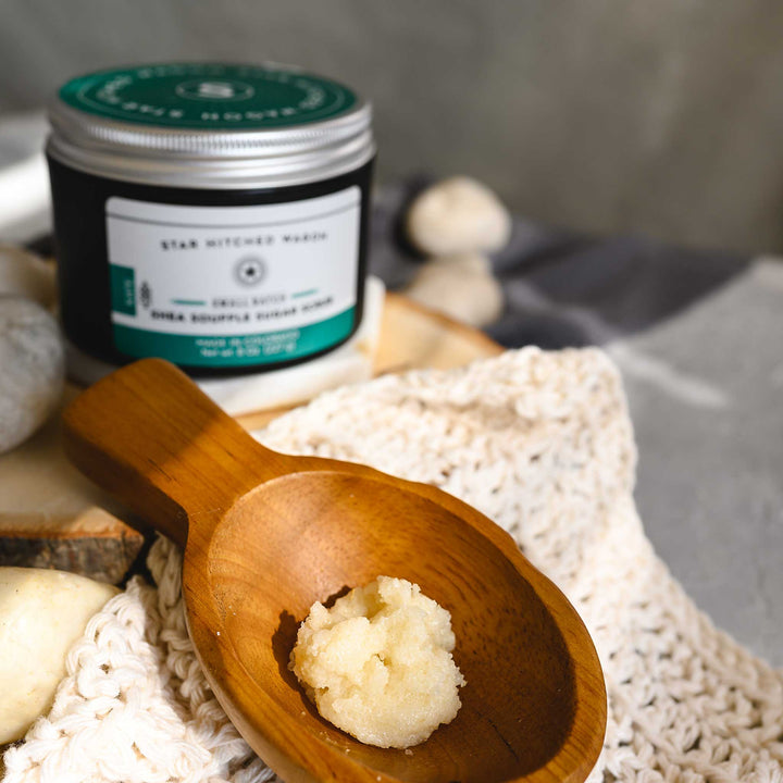 An 8 ounce container of Kate shea soufflé  sugar scrub is displayed with a wooden spoon showing the product, surrounded by natural elements like rocks and a knit washcloth, on a wooden surface.