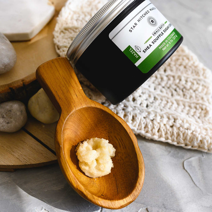 An 8 ounce jar of Hazel Shea Souffle Sugar Scrub is displayed on a wooden surface with a wooden spoon resting on a knit cloth, surrounded by small river stones.