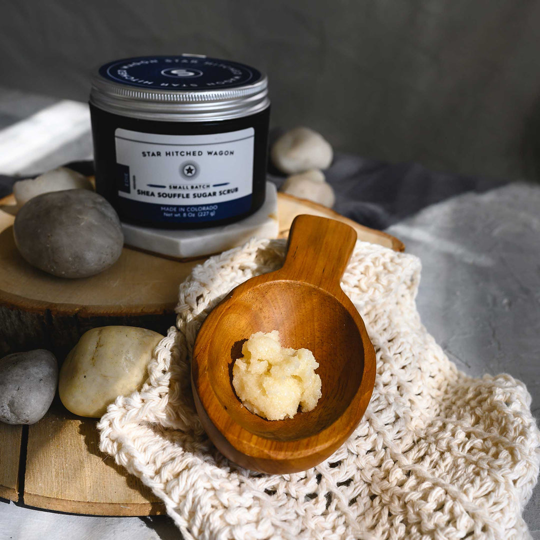 An 8 ounce jar of Edie Shea Souffle Sugar Scrub is displayed on a wooden surface, accompanied by a pebbles, wooden scoop and a knit wash cloth.