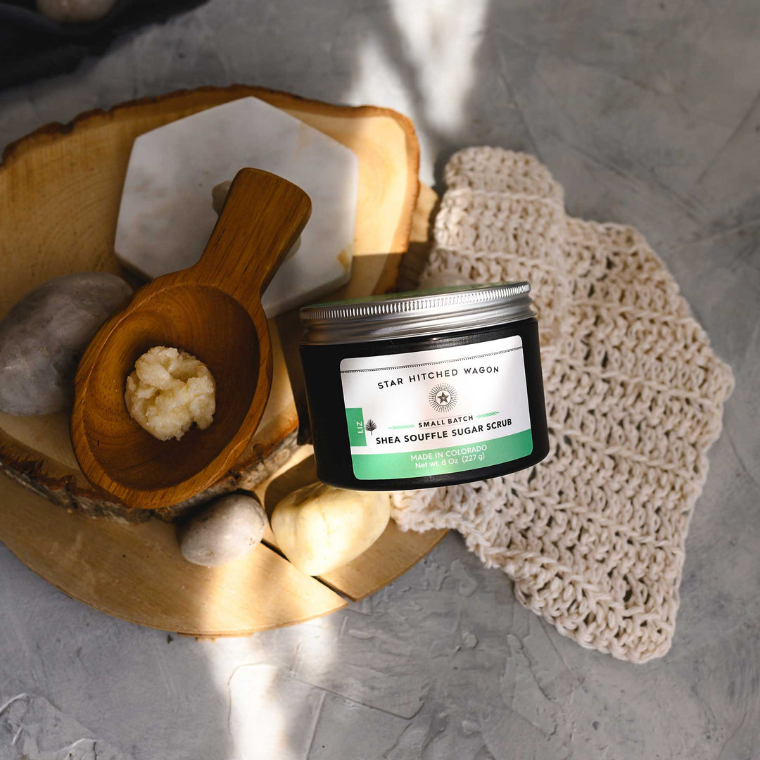 A jar of Liz 8 Ounce Shea Soufflé Sugar Scrub is displayed on a wood and stone surface with a wooden spoon, knit wash cloth and small river stones.