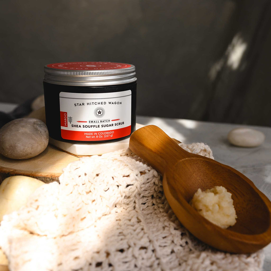 An 8 ounce jar of Margo Shea Soufflé Sugar Scrub is displayed on a wooden surface, accompanied by a pebbles, wooden scoop and a knit wash cloth.