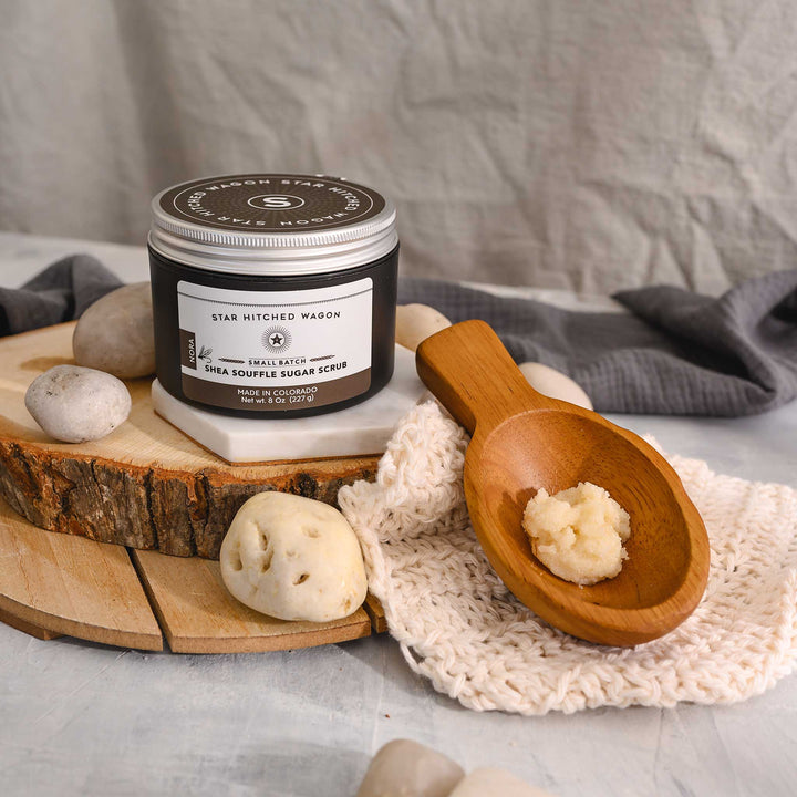 An 8 ounce jar of Nora Shea Soufflé Sugar Scrub is displayed on a wood and stone surface, accompanied by a pebbles, wooden scoop and a knit wash cloth.