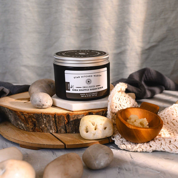 An 8 ounce jar of Nora Shea Soufflé Sugar Scrub is displayed on a wood and stone surface, accompanied by a pebbles, wooden scoop and a knit wash cloth.
