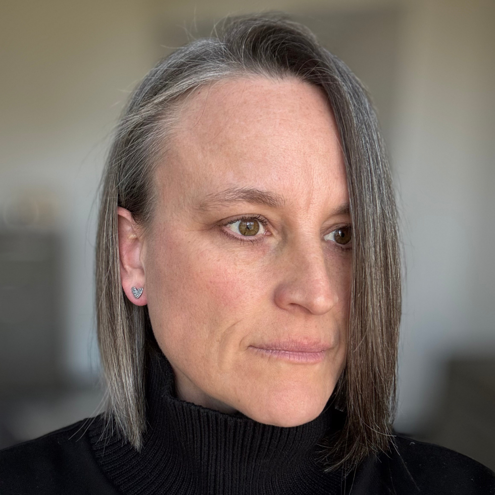 Woman with short dark hair wearing a black turtleneck sweater and small floral embossed sterling silver heart post earrings against a blurred background.
