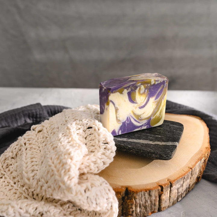 Jane essential oil soap bar with swirls of purple, green, and cream, resting on a stone and wooden surface next to a cream colored knit washcloth with a grey towel in the background.