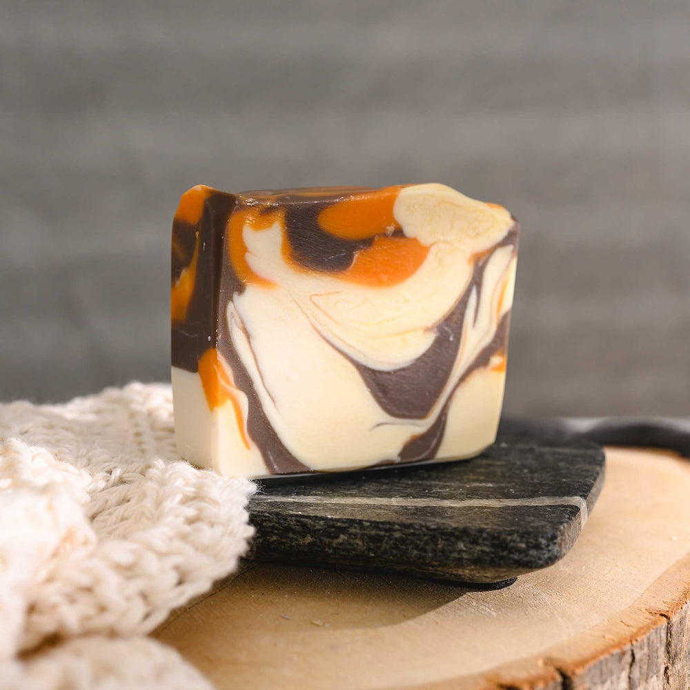 Jess essential oil soap bar with a swirl pattern of orange, brown, and cream colors, placed on a stone and wood surface with a cream colored knitted washcloth beside it.