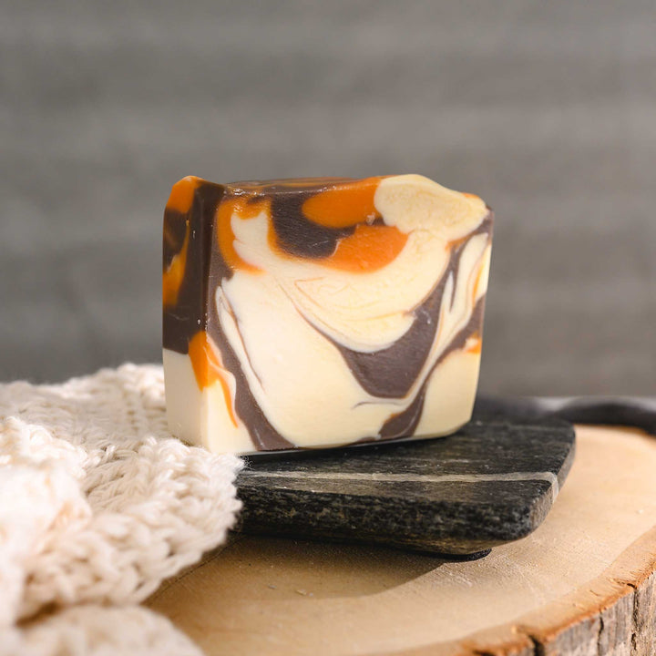 Jess essential oil soap bar with a swirl pattern of orange, brown, and cream colors, placed on a stone and wood surface with a cream colored knitted washcloth beside it.