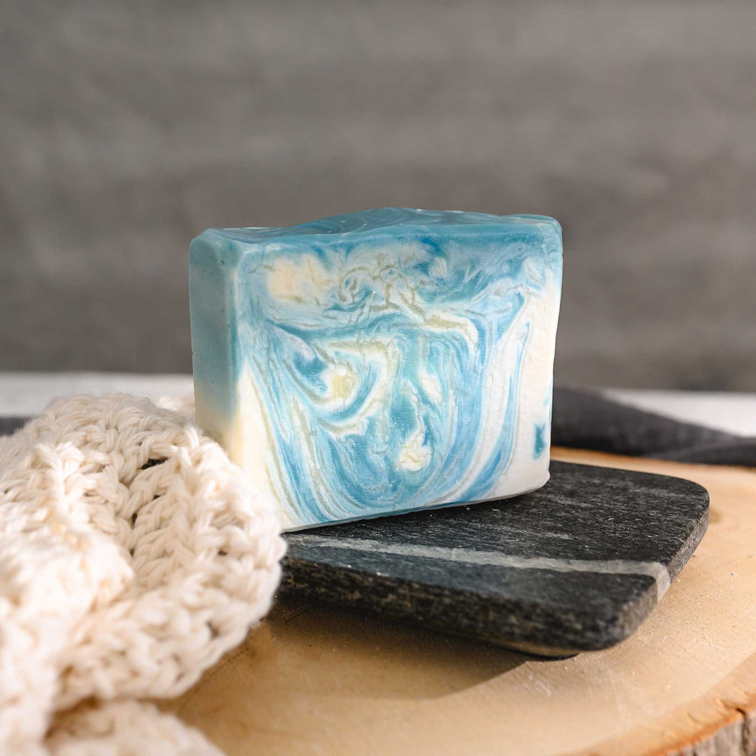 Kate essential oil soap bar with swirls of blue and white, placed on a stone and wood surface next to a cream colored knitted washcloth with a grey towel in the background.