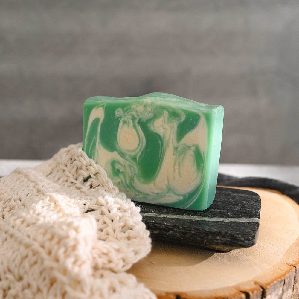 Liz essential oil soap bar with swirls of pale green and white, resting on a stone and wood surface with a cream colored washcloth beside it and a grey towel in the background.