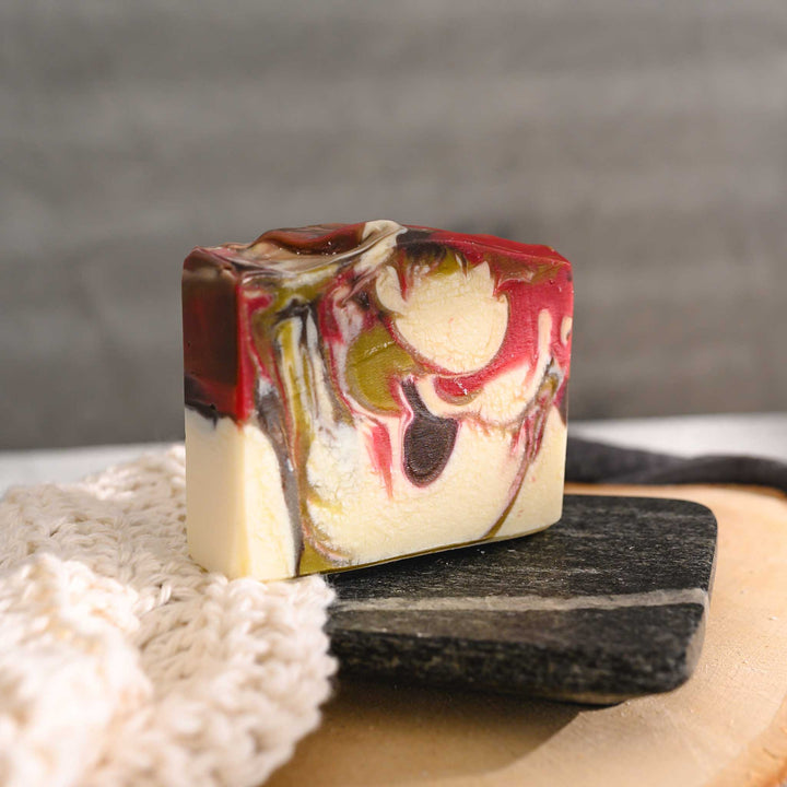 Margo essential oil soap bar with swirls of green, red, brown and white, resting on a stone and wood surface next to a cream colored washcloth with a grey towel in the background.