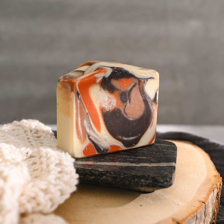 Nora essential oil soap bar with a swirl pattern in shades of orange, brown, and white, placed on a stone and wooden surface next to a crocheted washcloth with a grey towel in the background.