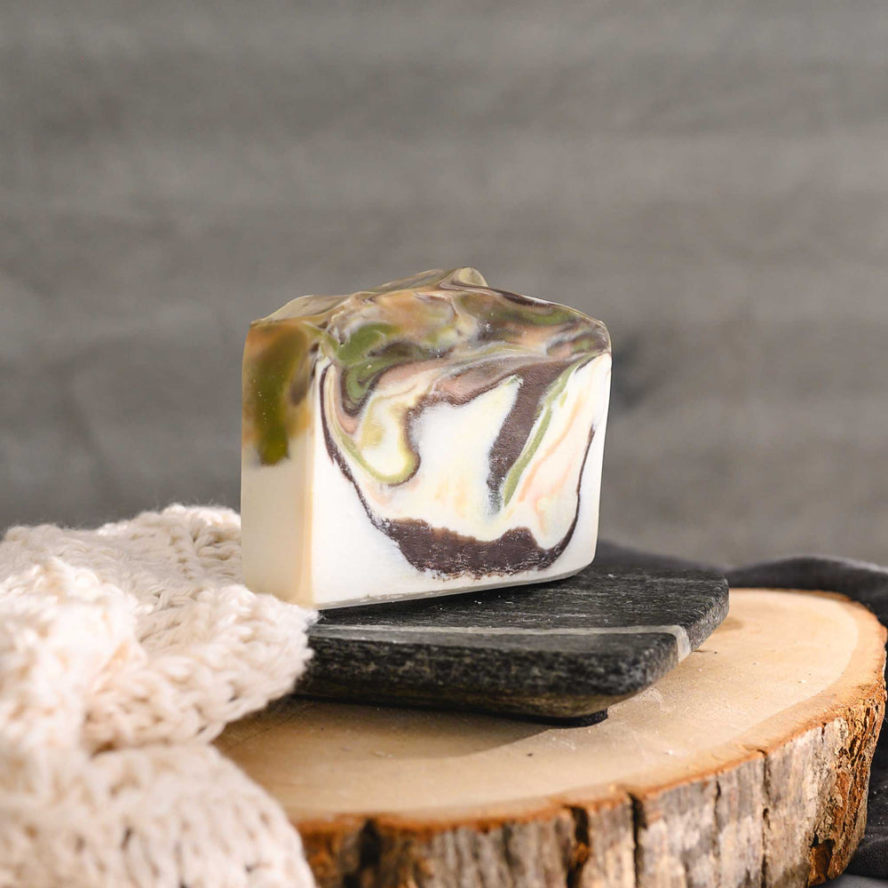 Hazel essential oil soap bar with swirls of peach, green, brown, and cream, placed on a wooden surface with a knit washcloth beside it.