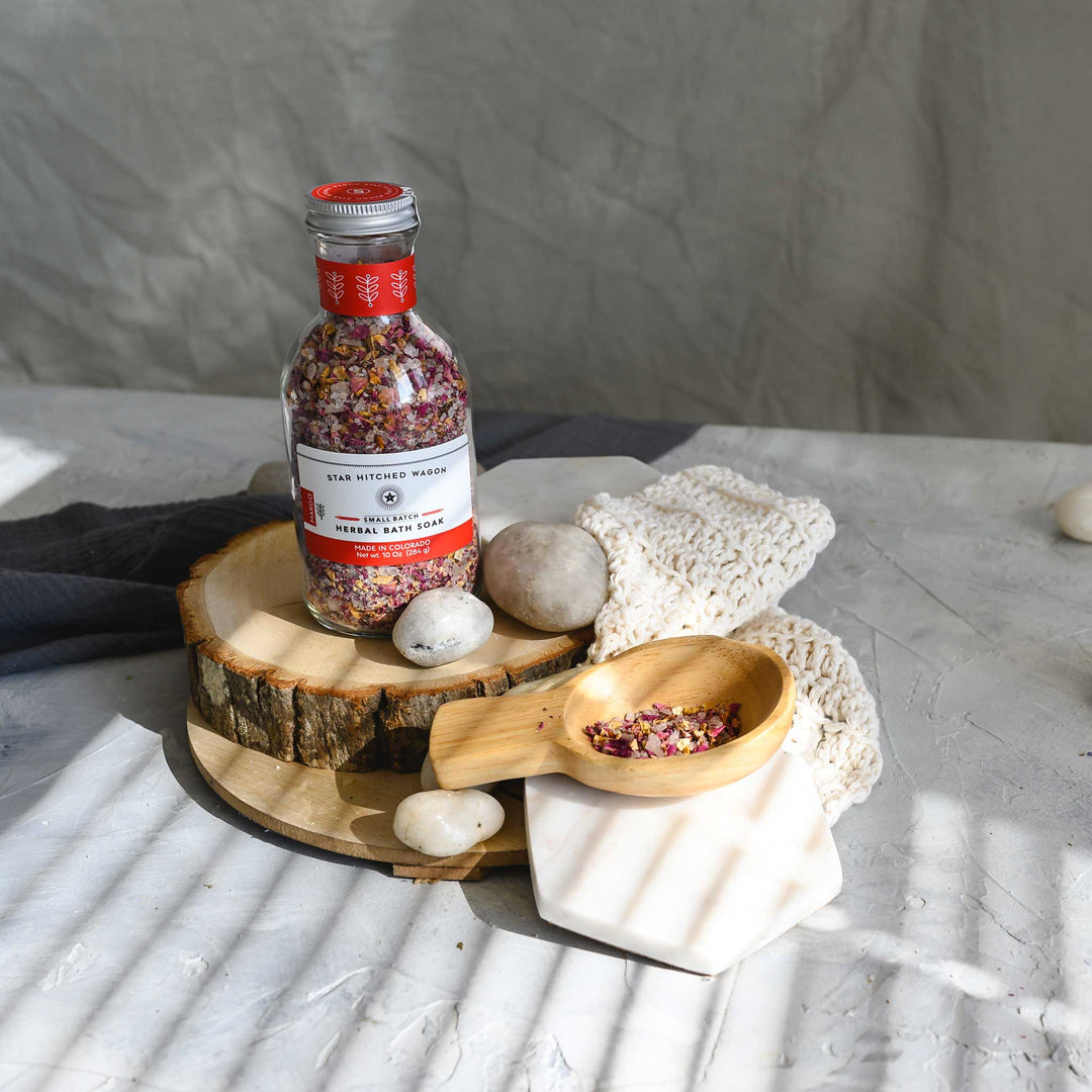 A glass bottle of Margo herbal bath soak with a screw top lid, placed on a knit washcloth next to a wooden scoop, on a textured grey surface, with white stones and a wooden surfaces in the background.