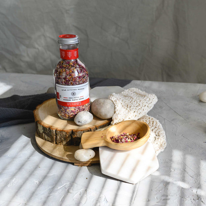 A glass bottle of Margo herbal bath soak with a screw top lid, placed on a knit washcloth next to a wooden scoop, on a textured grey surface, with white stones and a wooden surfaces in the background.