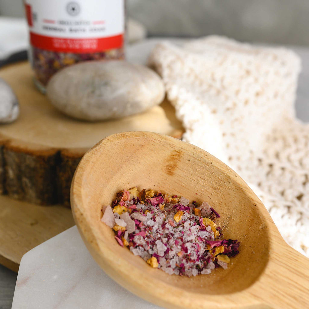 A close up of Margo herbal bath soak in a wooden scoop next to a knit washcloth, a wooden slab and small river stones.