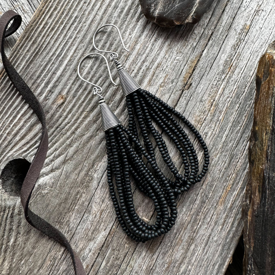 A pair of black seed bead earrings in loop style alongside a ribbon, displayed on a wooden surface.