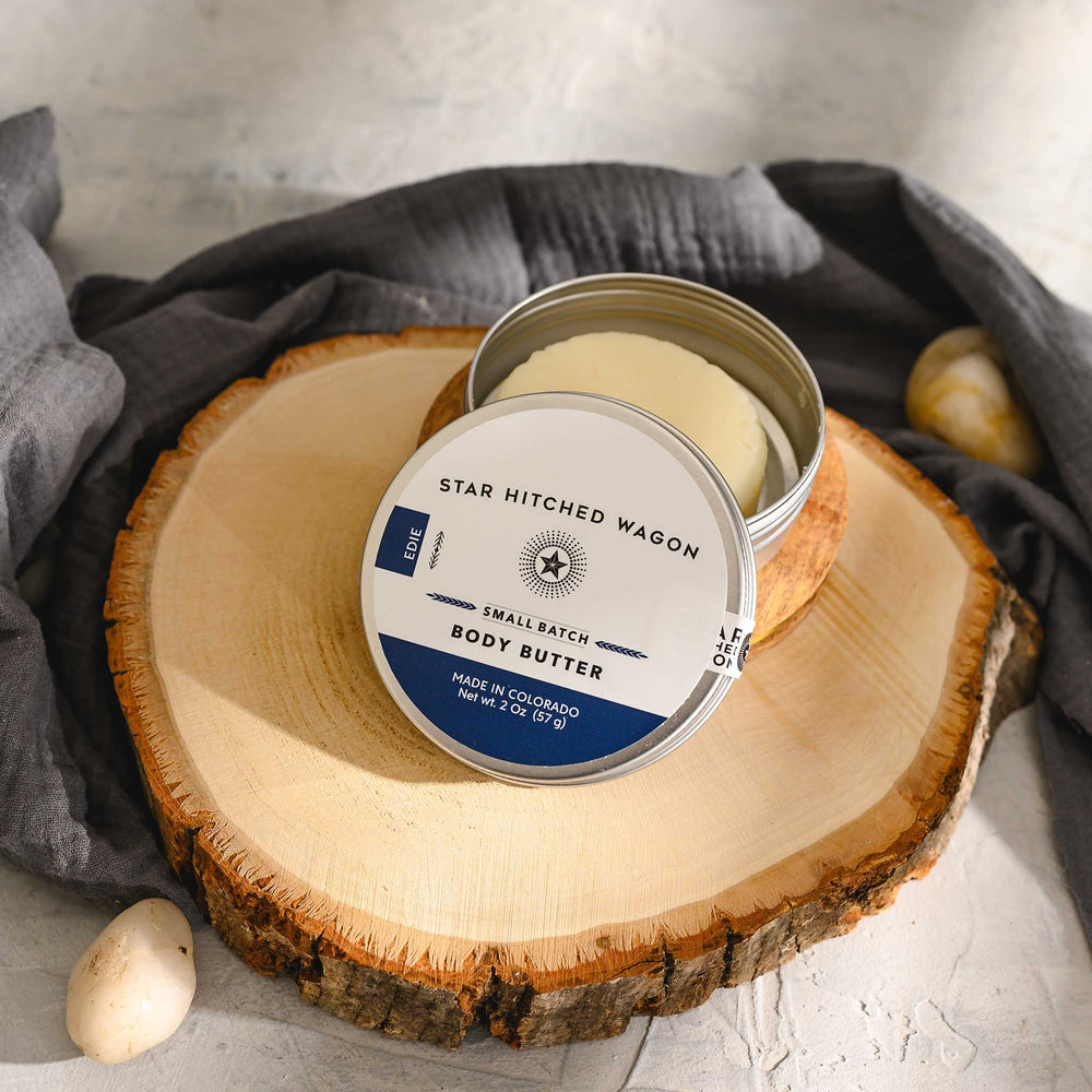 Edie 2 Ounce Body Butter Moisturizer on wooden slab with pebbles and grey towel in background.