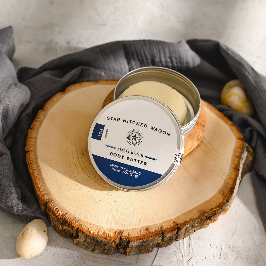 Edie 2 Ounce Body Butter Moisturizer on wooden slab with pebbles and grey towel in background.
