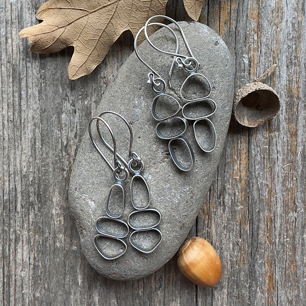 Two pairs of sterling silver cairn earrings on river stone and wood background with acorn and leaf accents.