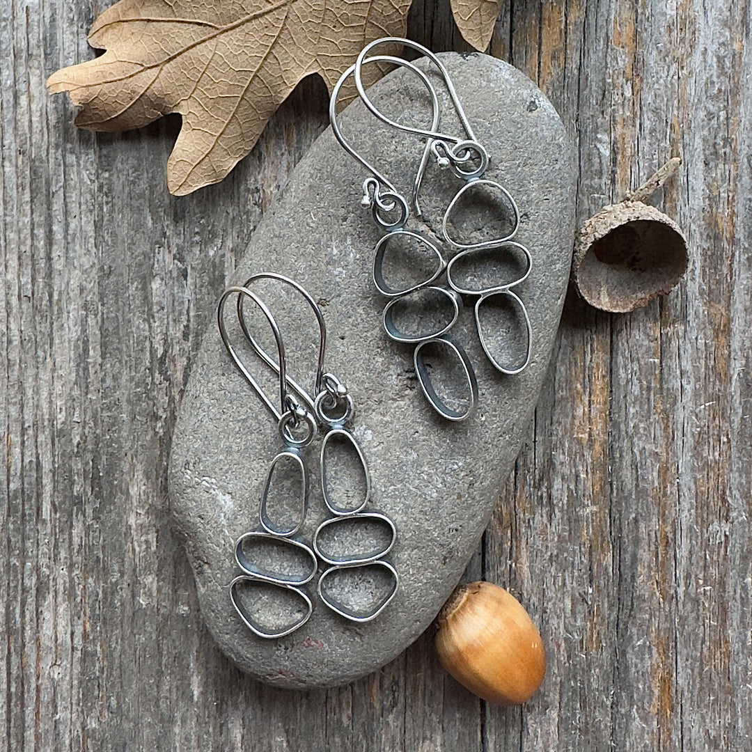 Two pairs of sterling silver cairn earrings on river stone and wood background with acorn and leaf accents.