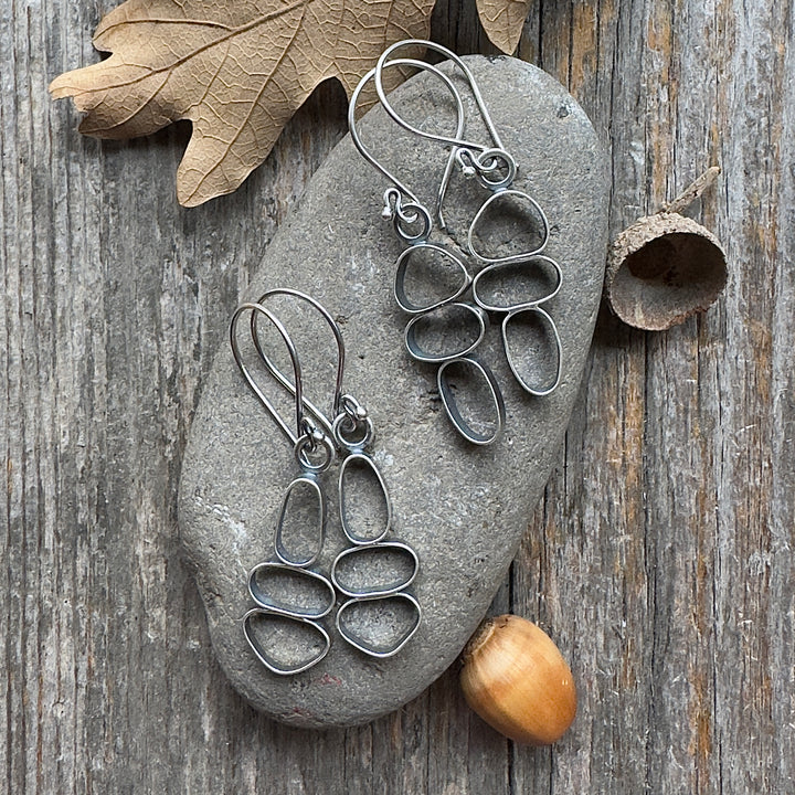 Two pairs of sterling silver cairn earrings on river stone and wood background with acorn and leaf accents.