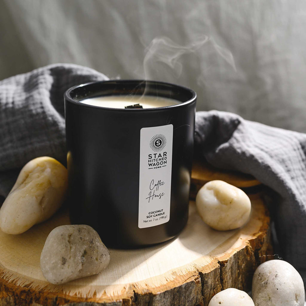 Coffee House Candle with wooden wick just blown out in a matte black jar with label. Linen towel, rocks and wood log surround candle.