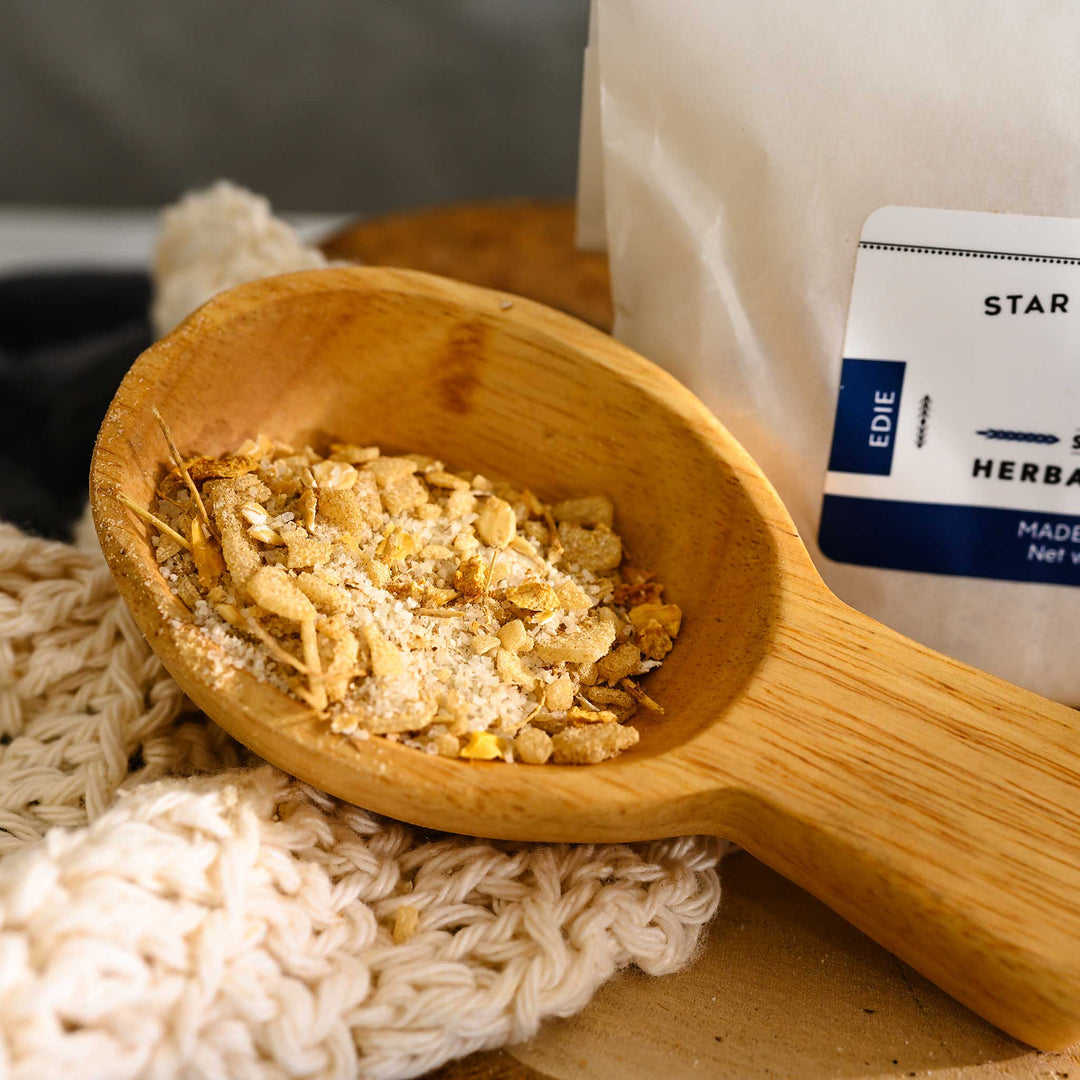 Wooden scoop filled with Edie Herbal Bath Soak next to full pouch on wooden slab next to a cream colored knit washcloth.