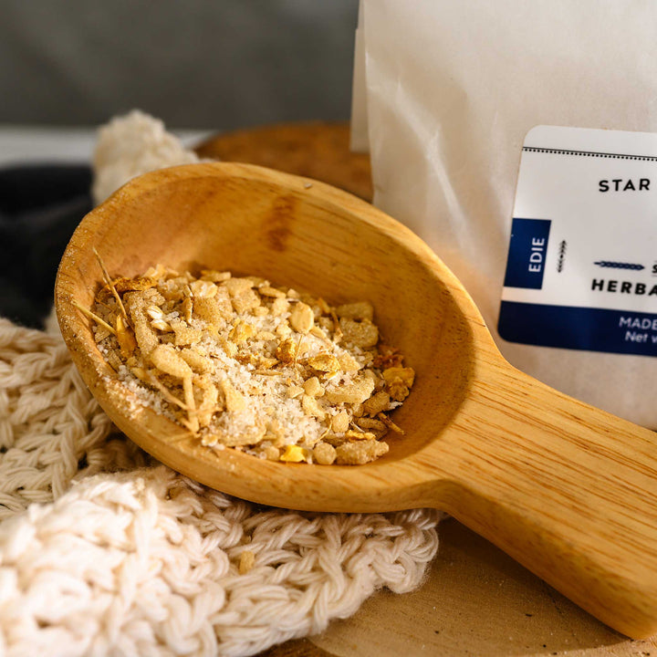 Wooden scoop filled with Edie Herbal Bath Soak next to full pouch on wooden slab next to a cream colored knit washcloth.
