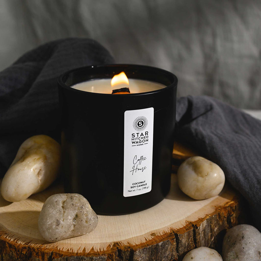 Coffee House Candle with wooden wick lit in a matte black jar with label. Linen towel, rocks and wood log surround candle.