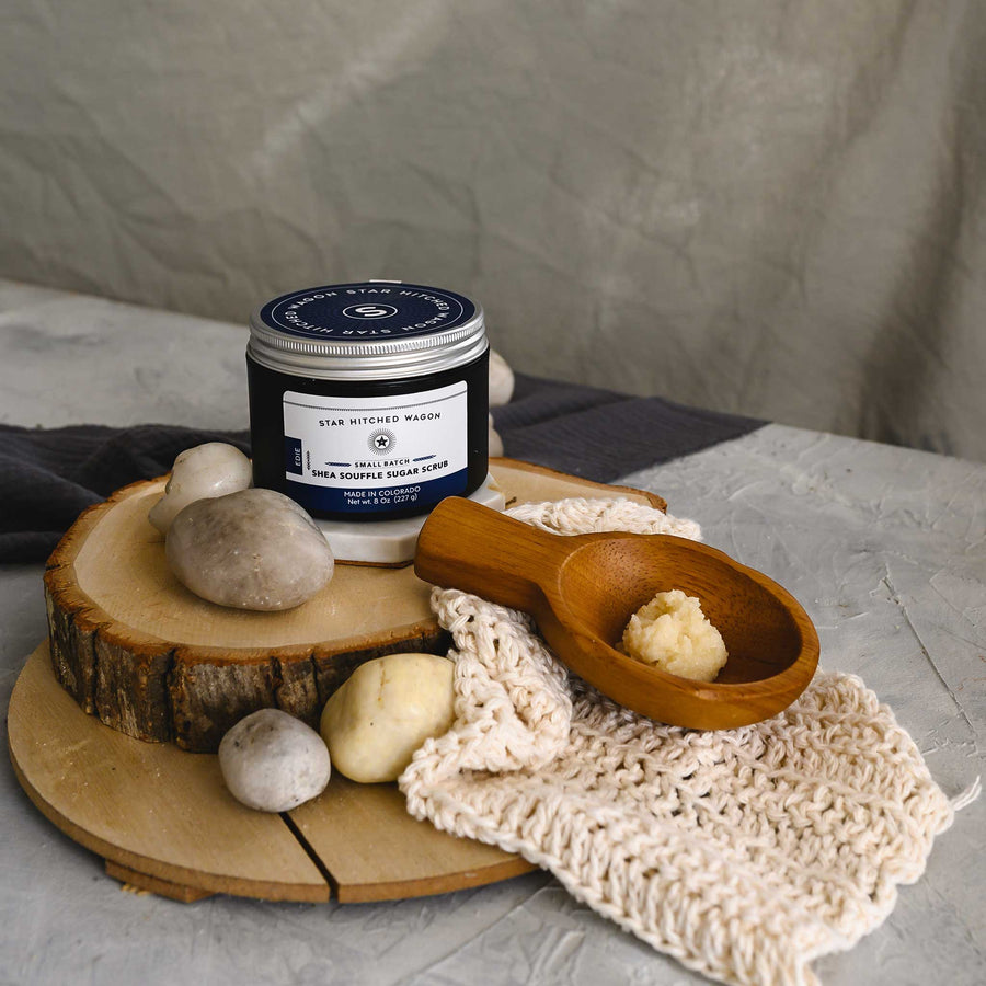 An 8 ounce jar of Edie Shea Souffle Sugar Scrub is displayed on a wooden surface, accompanied by a pebbles, wooden scoop and a knit wash cloth.