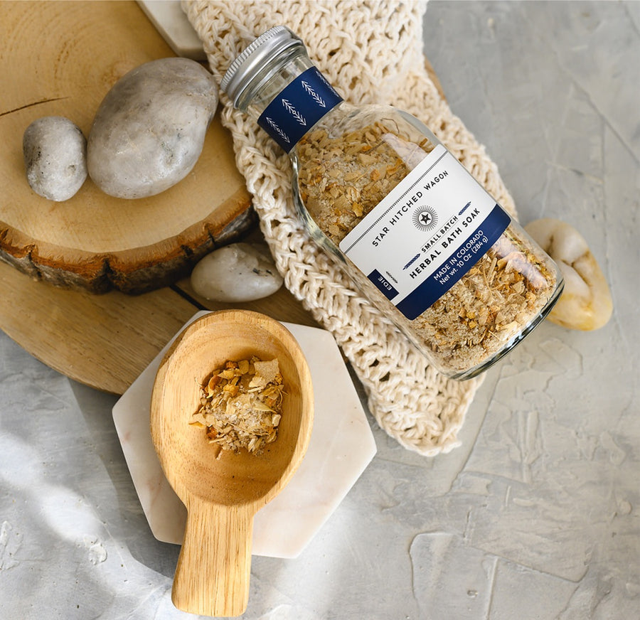 A glass bottle of Edie herbal bath soak with a screw top lid, placed on a knit washcloth next to a wooden scoop, on a textured grey surface, with white stones and a wooden surfaces in the background.