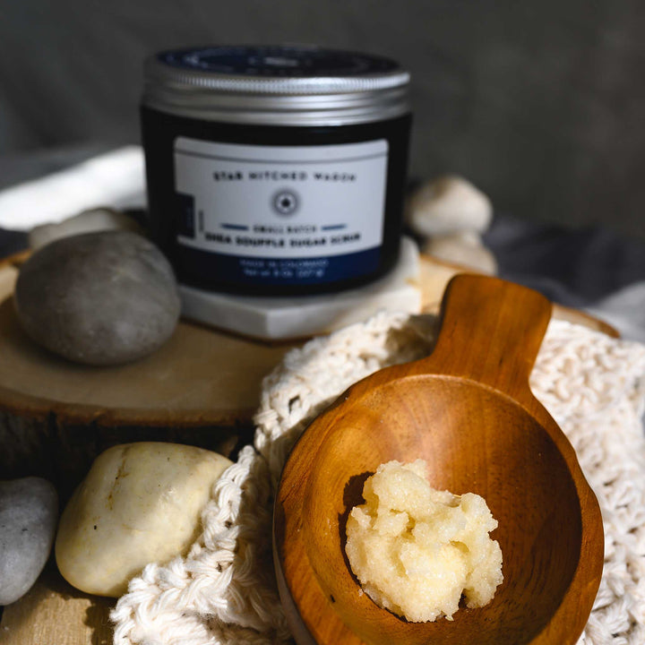 A close up of Edie Shea Souffle Sugar Scrub displayed in a wooden surface, accompanied by the full jar of scrub and a knit wash cloth.