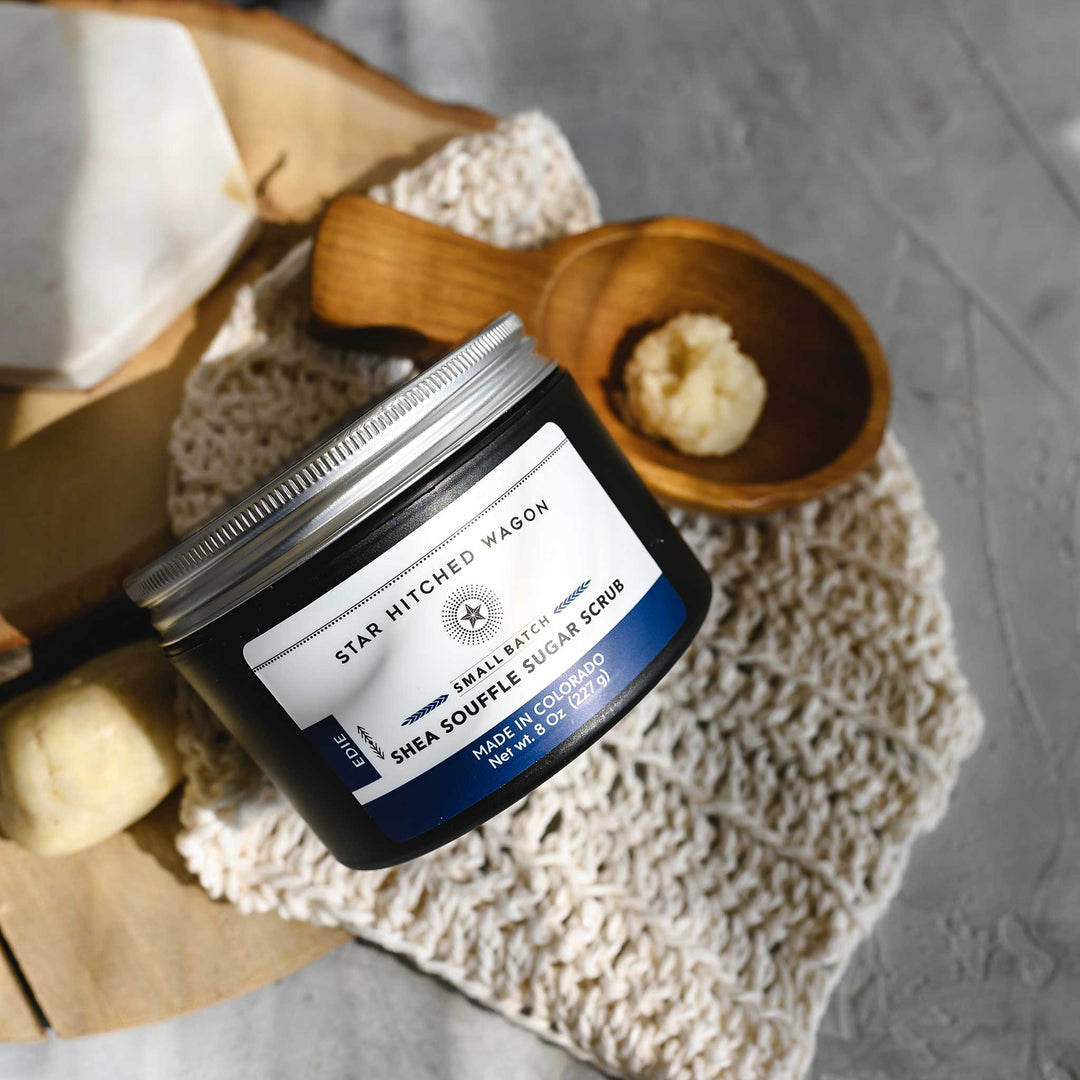 An 8 ounce jar of Edie Shea Souffle Sugar Scrub is displayed on a wooden surface, accompanied by a pebbles, wooden scoop and a knit wash cloth.