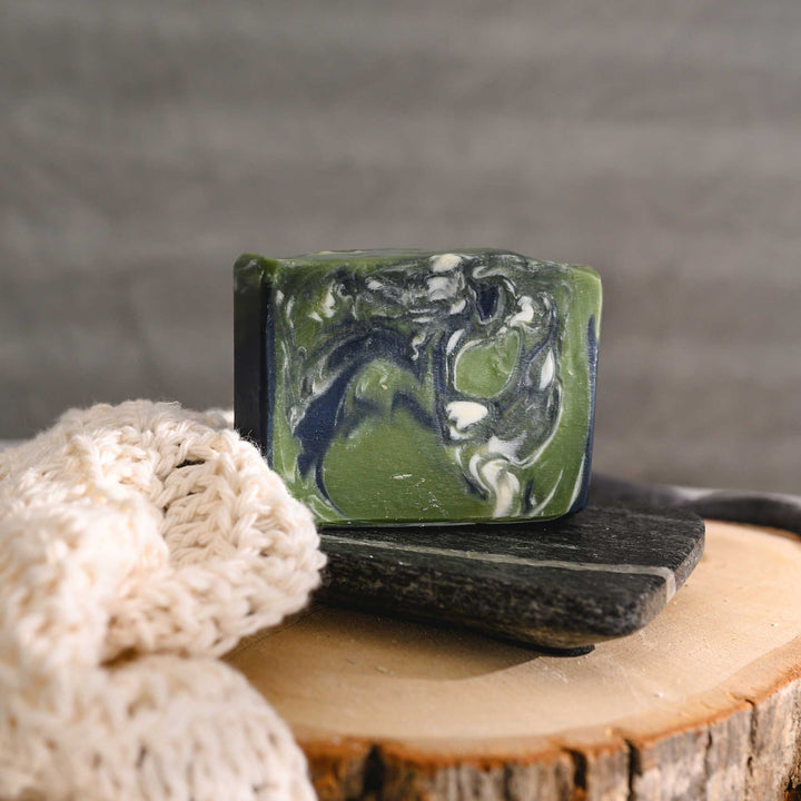 Edie essential oil soap bar with swirls of navy blue, green, and white, placed on a wooden surface with a knit washcloth beside it.