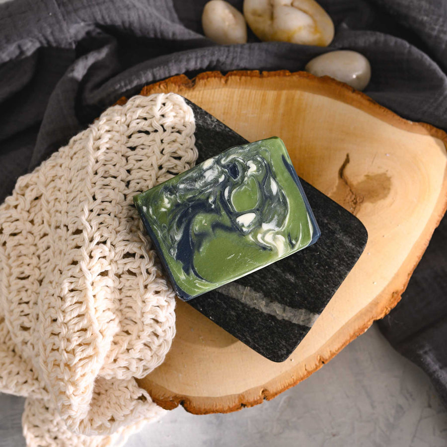 Edie essential oil soap bar with swirls of navy blue, green, and white, placed on a wooden surface with a knit washcloth beside it.